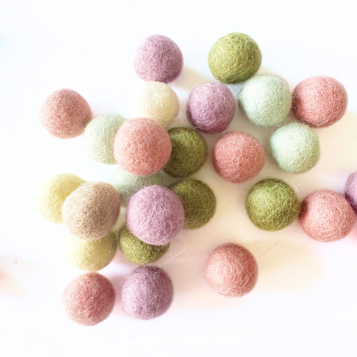 A cluster of felt wool balls in soft pastel colours including pink, purple, sage green, and pale yellow on a white background.