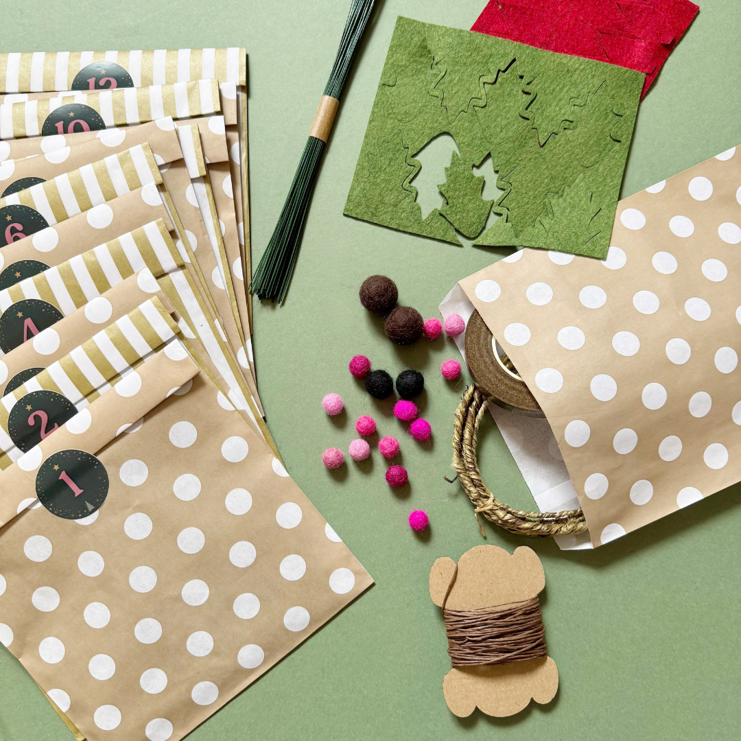 A flat-lay of paper bags and craft supplies on a sage green background. The main product shown is a set of kraft paper gift bags with white polka dots and alternating white and olive-green striped bands, each labelled with a circle containing a number. Also included are felt balls, florist wires, and felt sheets laser cut with shapes.