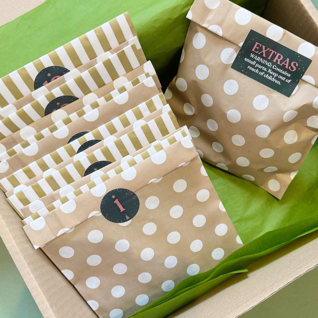 A box of gift bags and tissue paper. The bags are made of gold and white paper with white polka dots or stripes, each featuring a circular sticker with a number for 12 days of Advent.