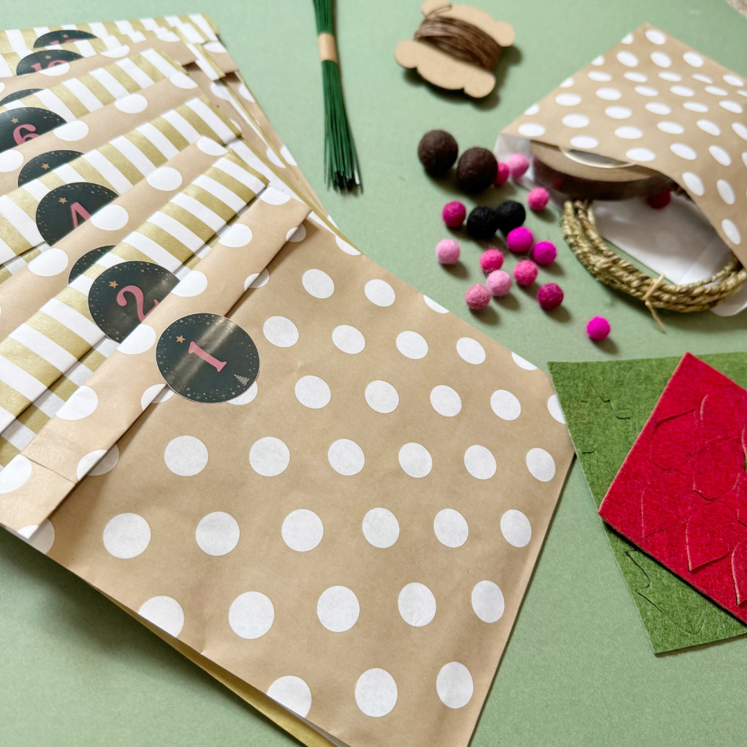 A flat-lay of paper bags and craft supplies on a sage green background. The main product shown is a set of kraft paper gift bags with white polka dots and alternating white and olive-green striped bands, each labelled with a circle containing a number. Also included are felt balls, florist wires, and felt sheets laser cut with shapes.