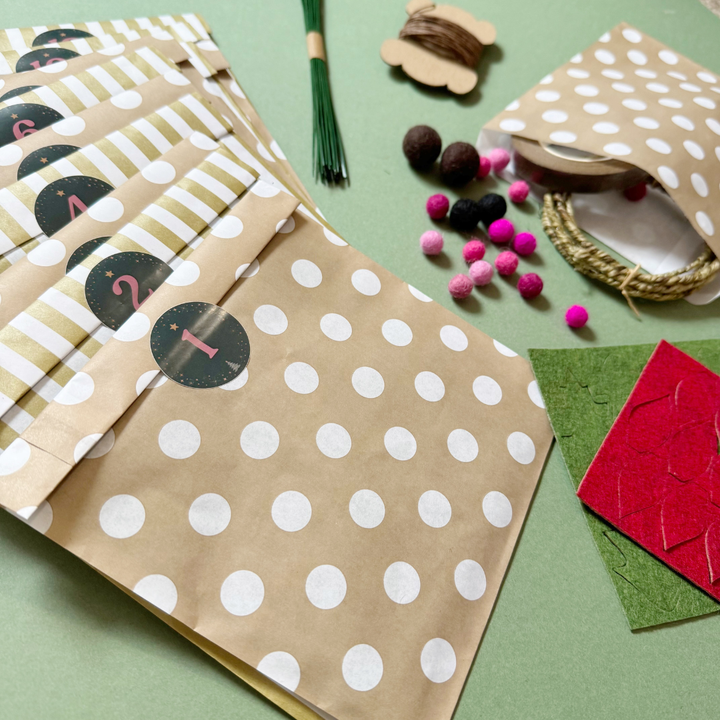 A flat-lay of paper bags and craft supplies on a sage green background. The main product shown is a set of kraft paper gift bags with white polka dots and alternating white and olive-green striped bands, each labelled with a circle containing a number. Also included are felt balls, florist wires, and felt sheets laser cut with shapes.