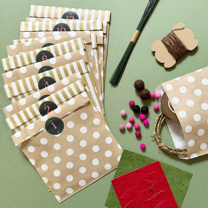 A flat-lay of paper bags and craft supplies on a sage green background. The main product shown is a set of kraft paper gift bags with white polka dots and alternating white and olive-green striped bands, each labelled with a circle containing a number. Also included are felt balls, florist wires, and felt sheets laser cut with shapes.
