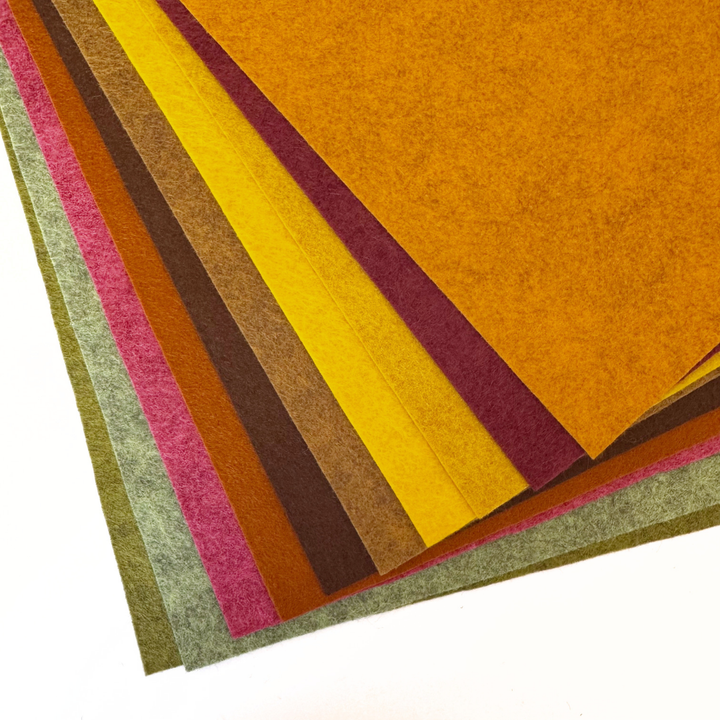 A stack of textured felt sheets fanned out diagonally, showing multiple colours including mustard yellow, orange, burgundy, yellow, brown, pink, and green on a white background.