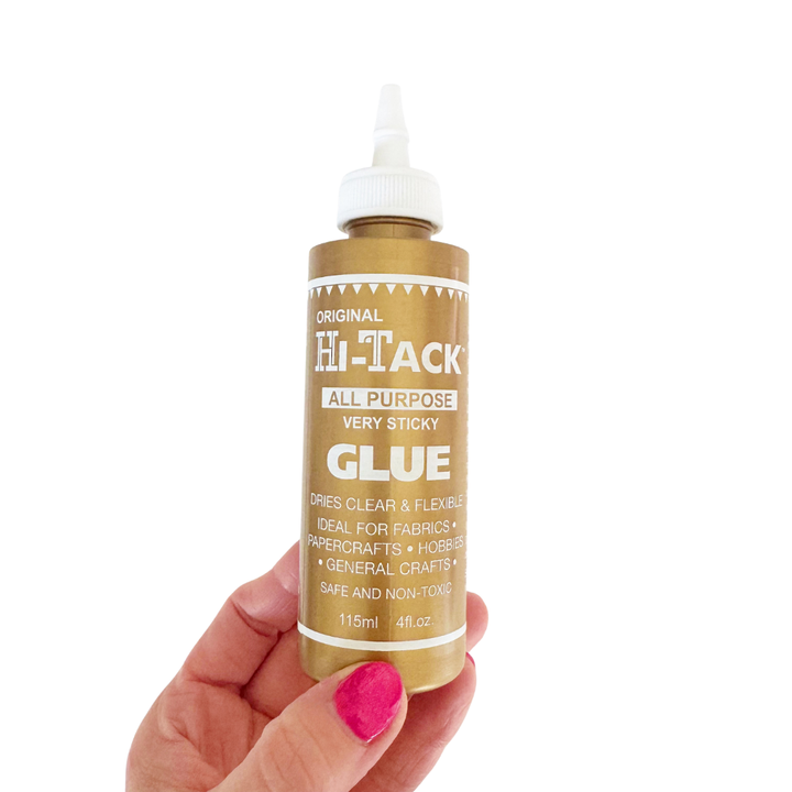 A hand with bright pink nail polish holds a bottle of Hi-Tack All Purpose Glue against a plain white background. The bottle is metallic gold with a white cap and white printed text.