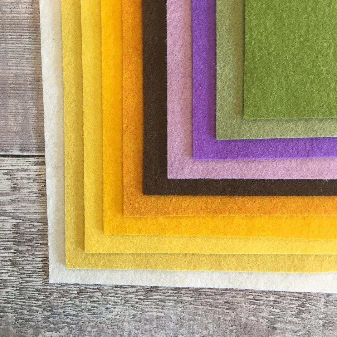 A stack of various colored felt sheets including white, yellow, gold, orange, brown, purple, blue, green, and gray, laid out on a wooden surface.