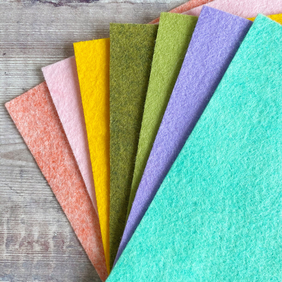 A collection of seven different colored felt sheets arranged in a fan-like spread on a wooden surface. Colors include coral, pink, gold, green, yellow, purple, and blue.