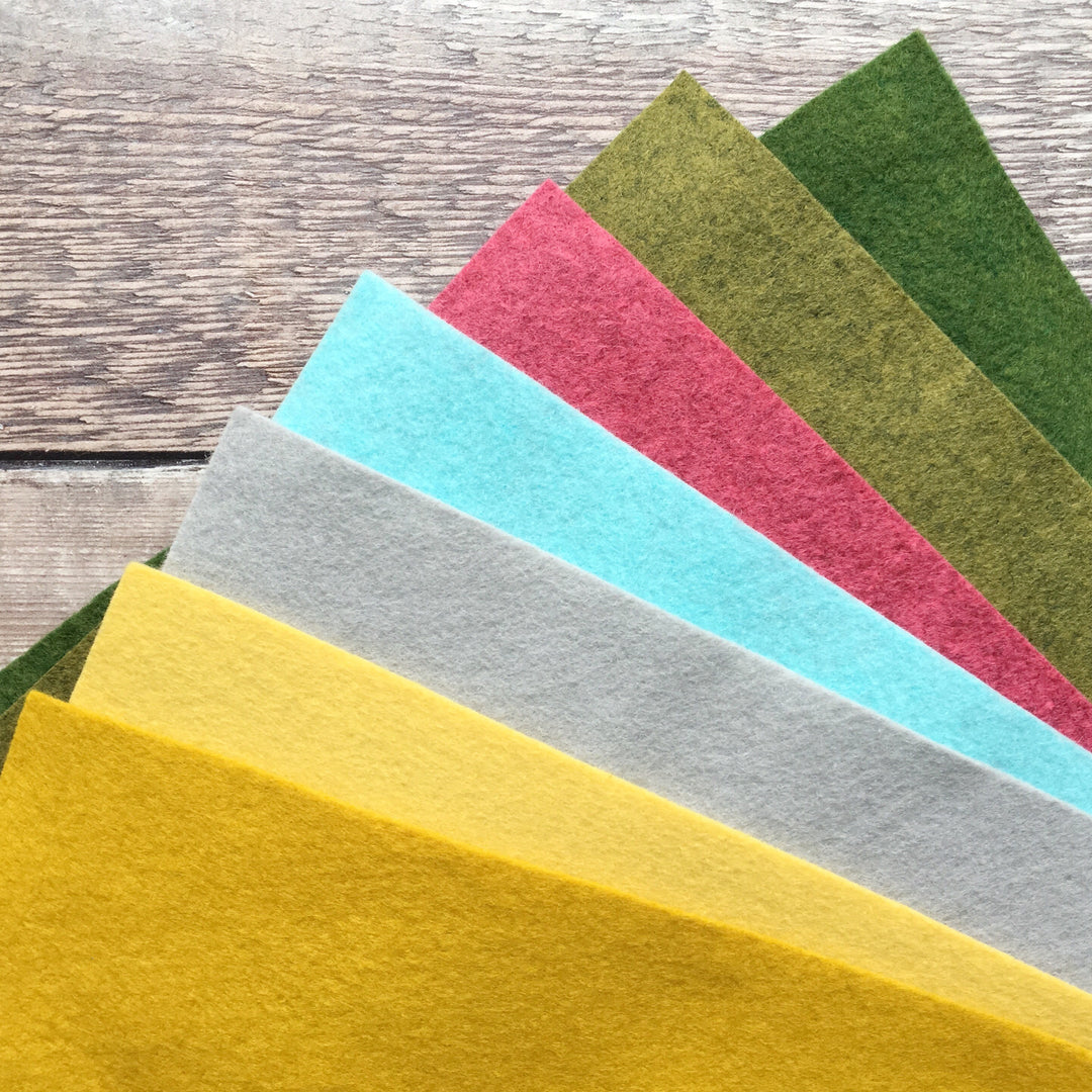 A collection of felt sheets in various colors including gold, yellow, silver, blue, pink, green, and grass green, laid out on a wooden surface.