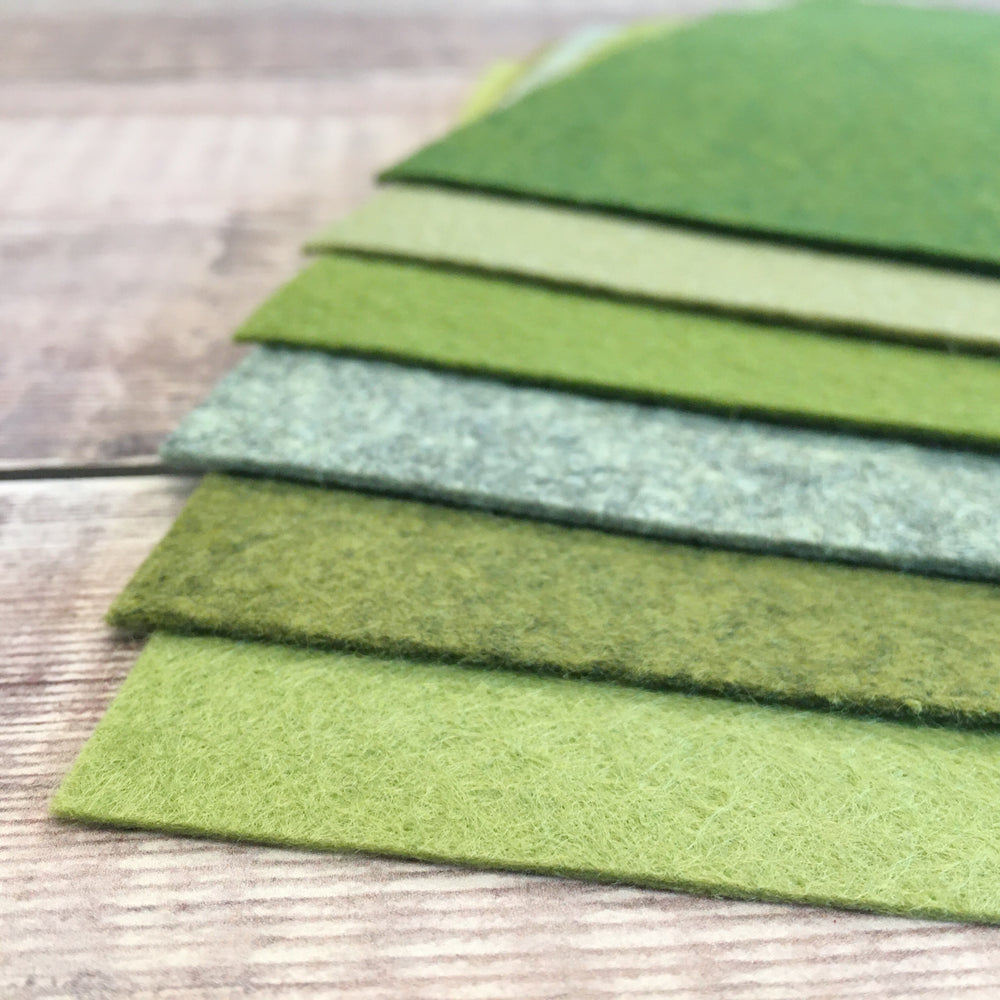 A selection of six green shades of wool-blend felt sheets, laid out on a wooden surface.
